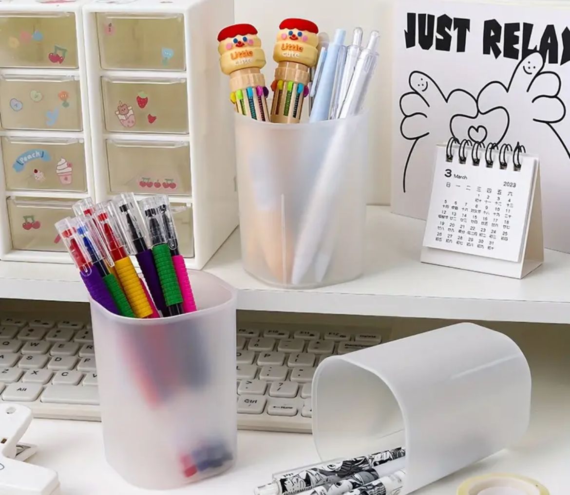 Office supplies including a pen holder with various pens, a highlighter, and a desk calendar, all displayed on a white desk with a 'Just Relax' sign in the background.